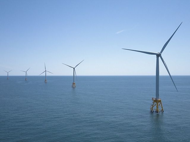 A view of five offshore wind energy turbines, with the right-hand turbine in the foreground stretching to the left-most turbine in the background. They sit on blue ocean water against a blue sky.